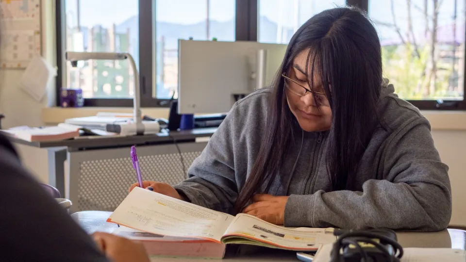 A student wearing glasses with long brown hair sits at a desk and looks at an open textbook.