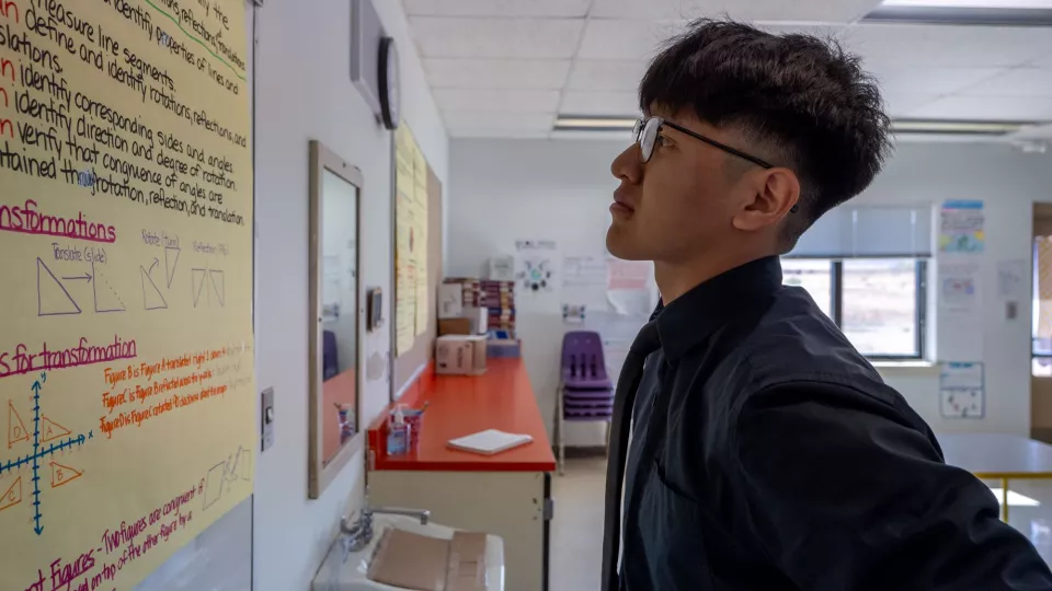 A student wearing glasses stands and reads a poster in a classroom.