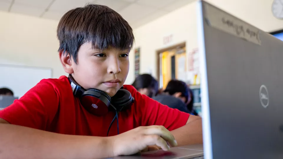 A young male student with brown hair sits at a desk in a classroom setting and looks at his computer.