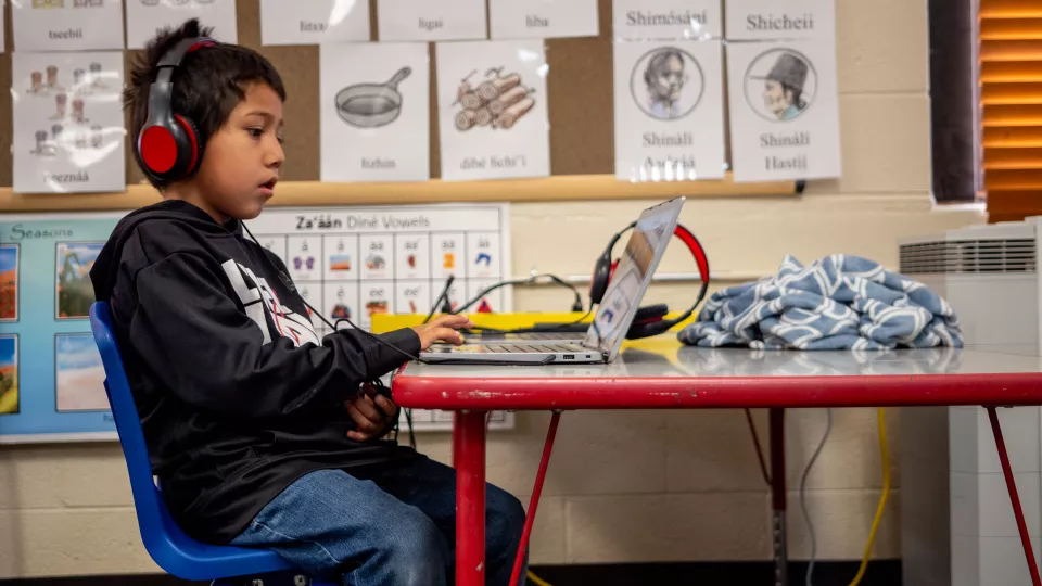 A young student sits at a desk in front of a laptop and wearing headphones.
