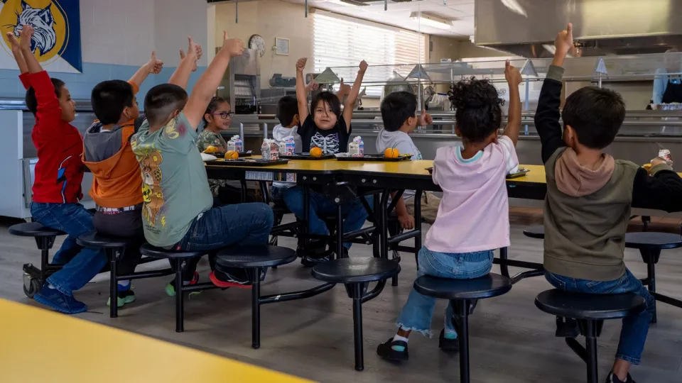 Young students sit at a cafeteria table with food in front of them, all giving thumbs up.