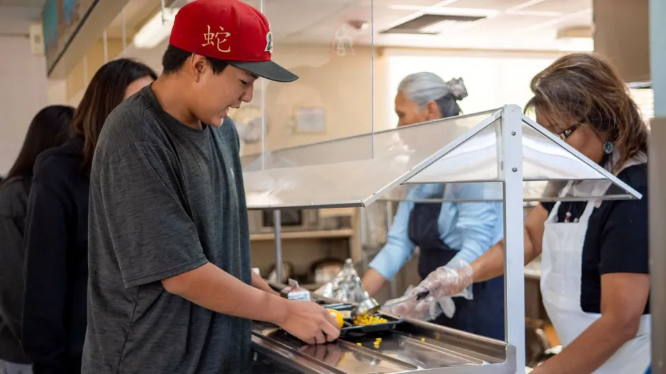A student in a gray shirt and red ball cap goes through the lunch line in a school cafeteria.