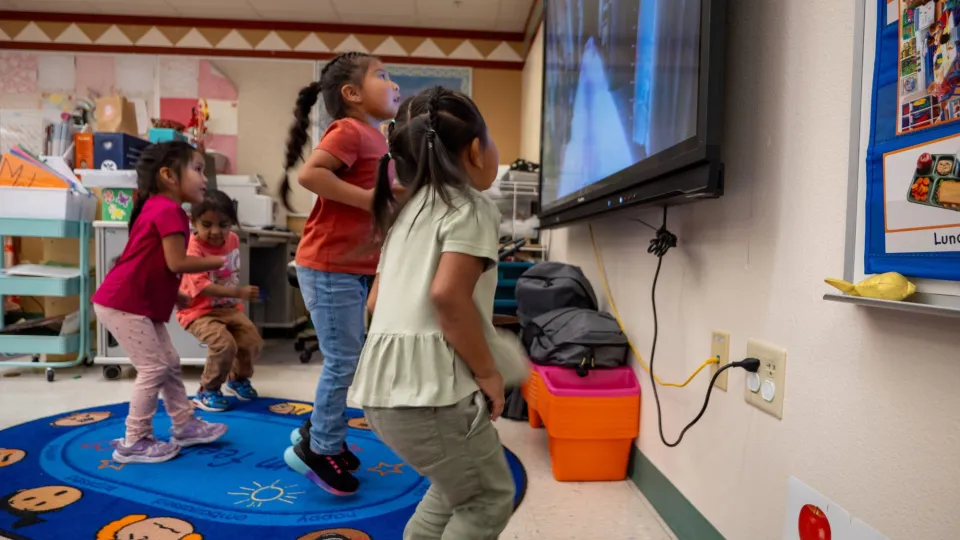 Young students stand on a rug in front of a classroom TV screen and jump along with a video.