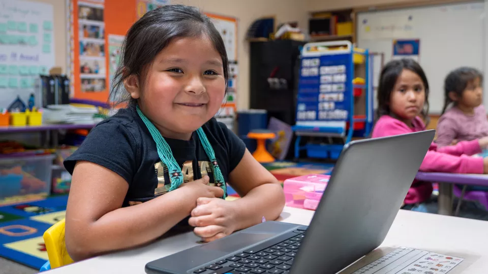 A elementary school student smiles while sitting at her computer at her desk.