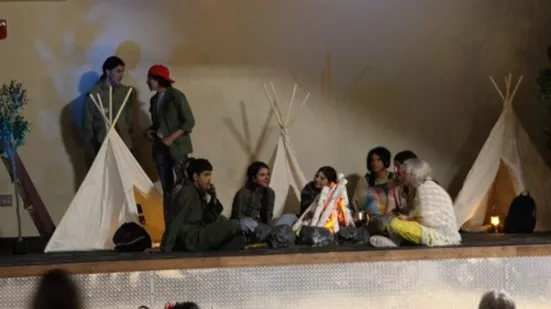 A group of students perform a scene from a play while sitting in a circle on the stage.