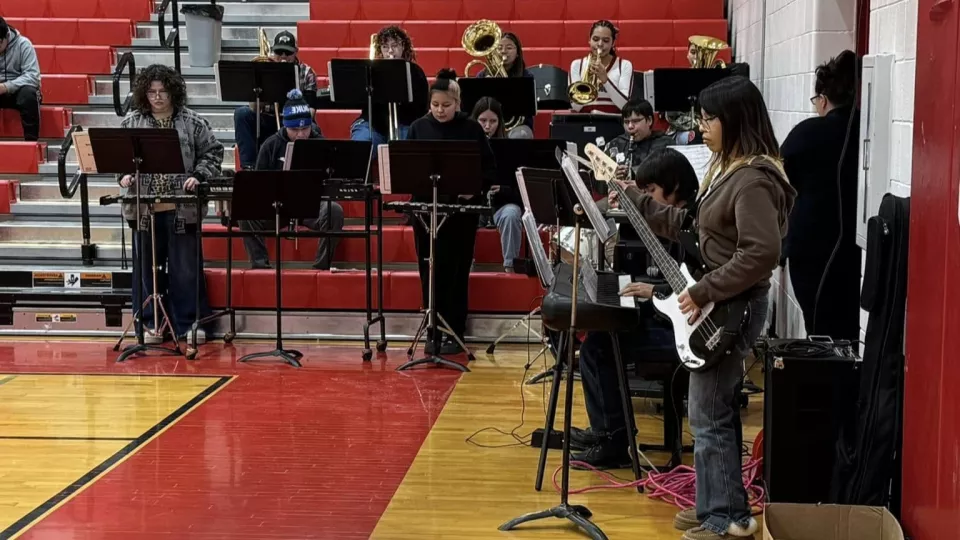 A high school band stands in the corner of a gymnasium and plays at an assembly.