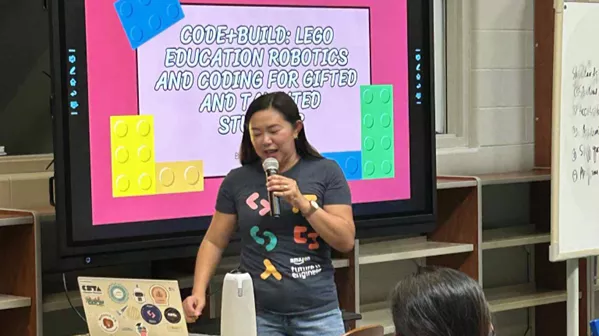 A teacher gives a presentation while standing at the front of a room, holding a microphone in front of a large monitor.