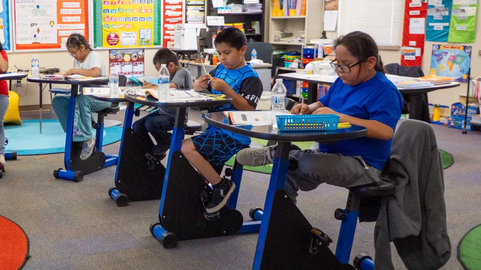 A group of students sit at their desks with stationary bikes.