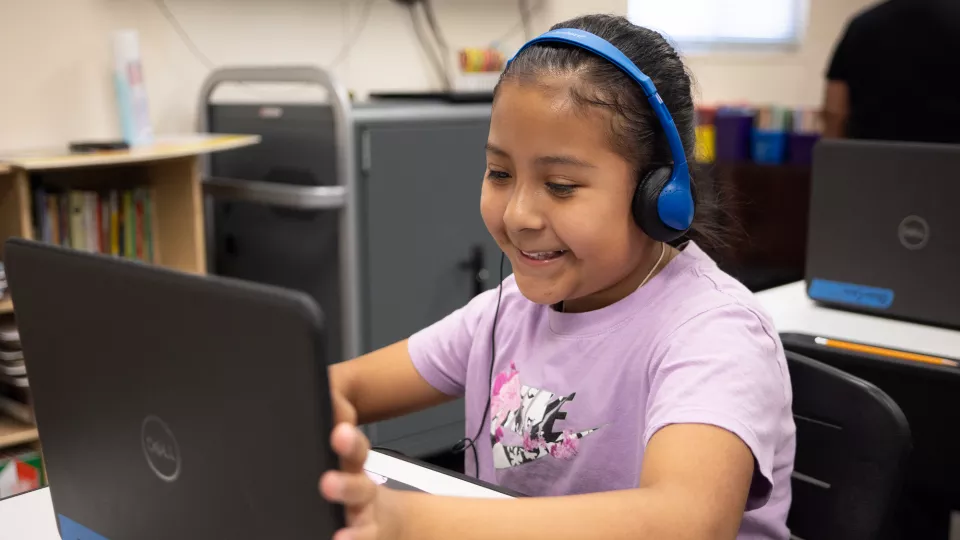 A young student sits at her desk in front of her laptop, smiling and wearing headphones.