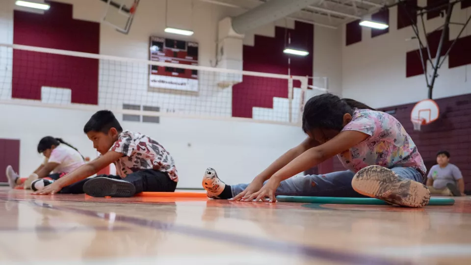 Students sit in a line on a gym floor and stretch.