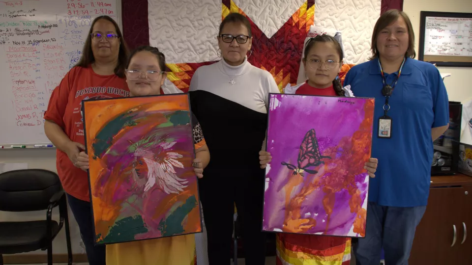 Two young female students hold up their paintings while three women stand behind them, posing for a photo.