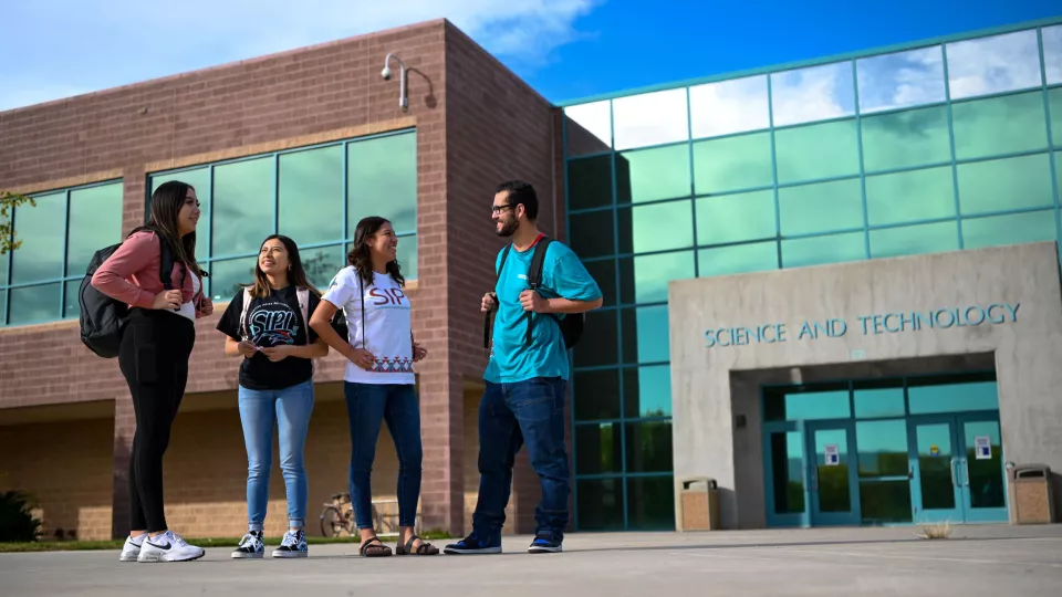 Four students wearing backpacks stand in front of a college campus building and talk.