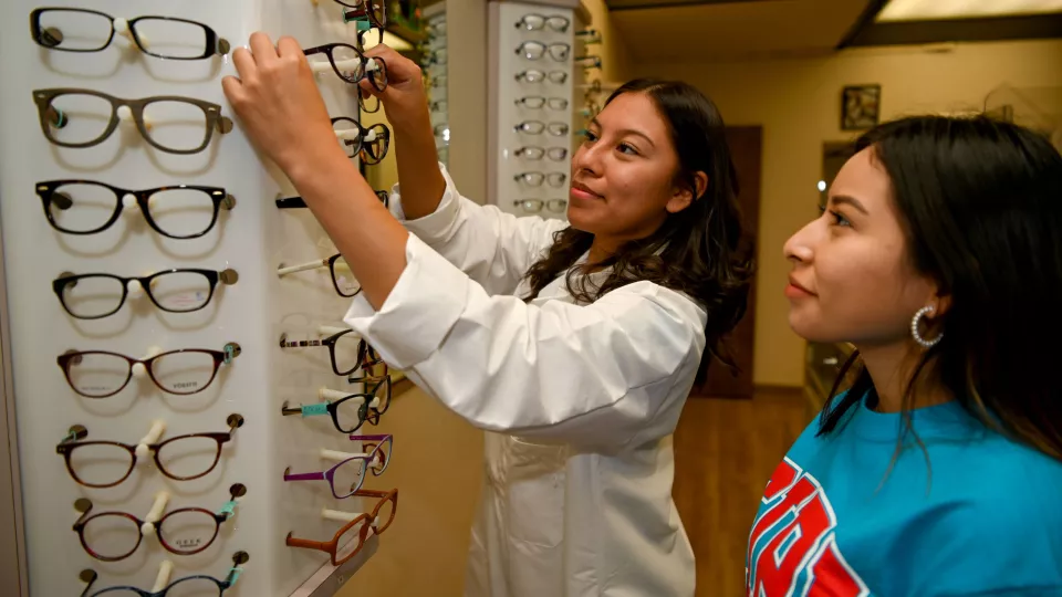 A young vision care program student helps another pick out glasses at a display.