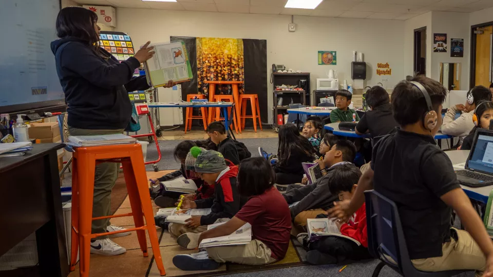 Young students sit on the ground with their books open in their laps in a classroom while looking at their teacher standing at the front of the room.