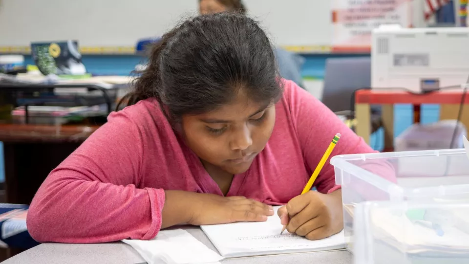 A young student in a pink sweater sits at her desk and writes with a pencil and paper.