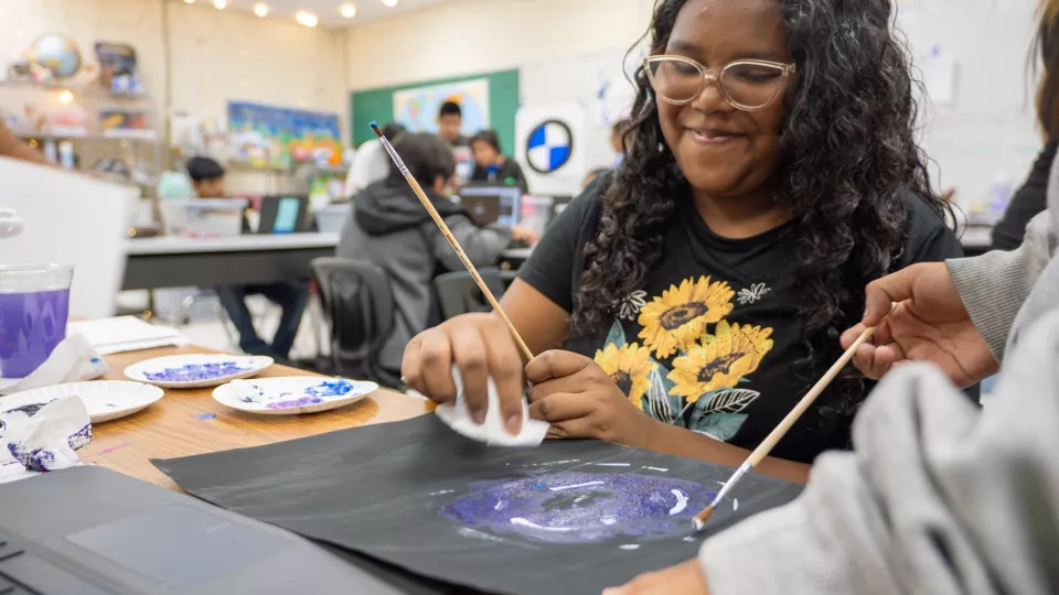 Two students sit at a table with art supplies and work together on a painting.
