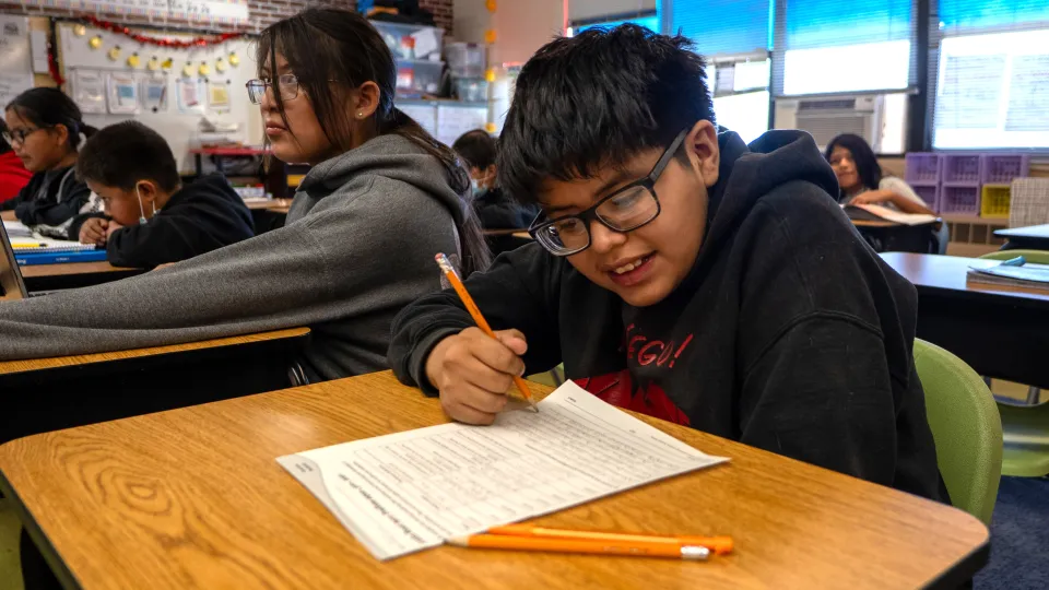 A student in a black hoodie sits at a desk in a classroom and writes on a worksheet with a pencil.