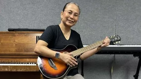 A woman in a black t-shirt holds a guitar while sitting down.