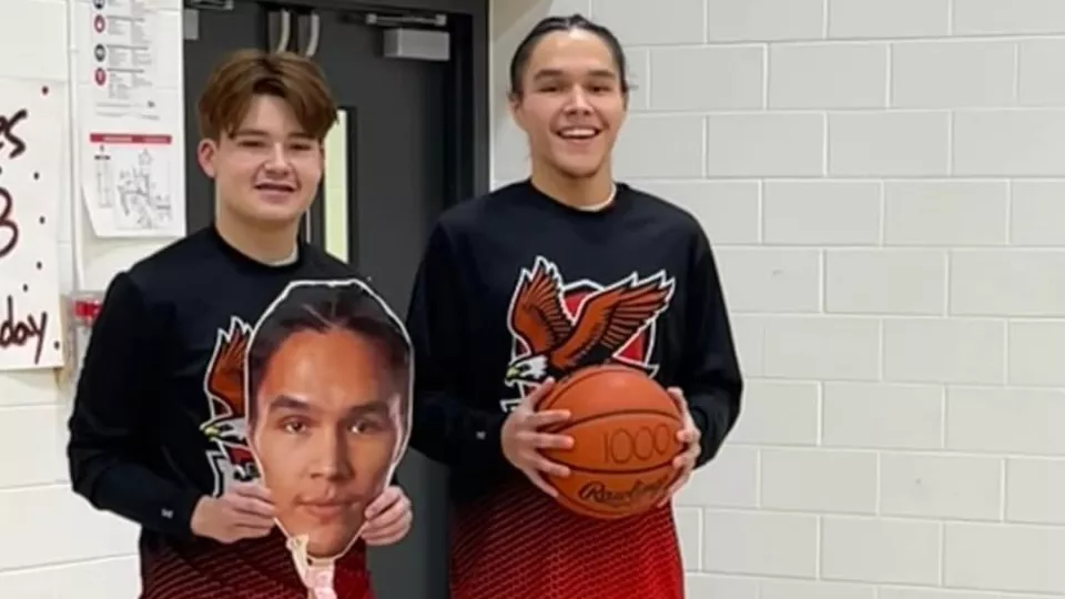 Two basketball players wearing matching team warm-up shirts stand in a gym, smiling. One holds a basketball marked "1000" while the other holds a large cutout of a face, celebrating a milestone achievement.
