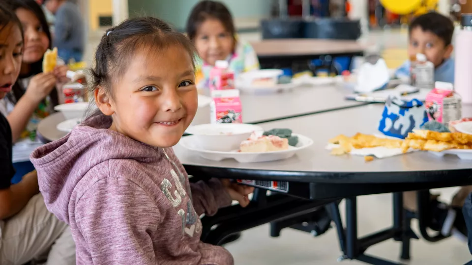 An elementary school student sits at a lunch table in a cafeteria and smiles.