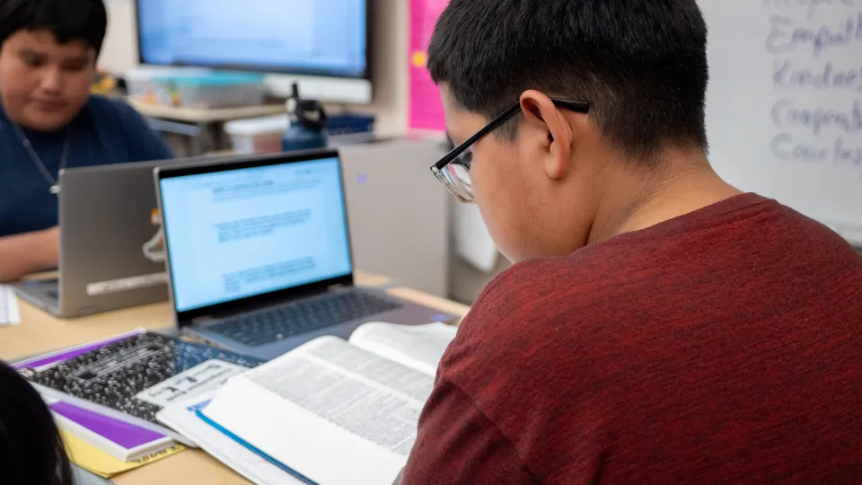 A student sits at a desk and looks at a textbook and his laptop.
