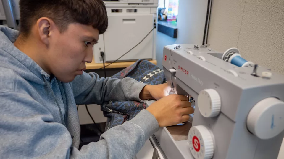A student in a grey hoodie sits at a sewing machine and sends a piece of fabric through it.