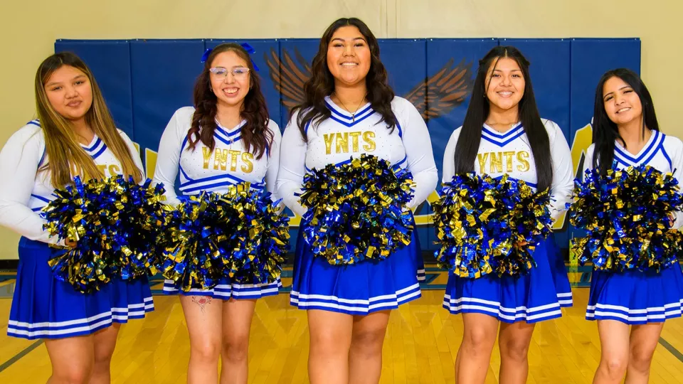 A group of five cheerleaders in white, blue and gold uniforms pose for a photo holding blue and gold pom poms.