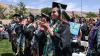 Students in black graduation robes and caps stand and clap in a celebratory outdoor setting.