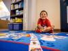 A young student in a red shirt and shoes sits on a brightly colored rug in a classroom and smiles.