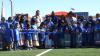 A large group of elementary school students and adults standing behind a blue ribbon that is being cut.